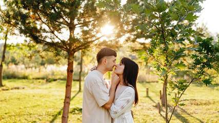 Naklejka premium Young Couple Kissing in Sunny Park at Golden Hour Surrounded by Trees and Greenery