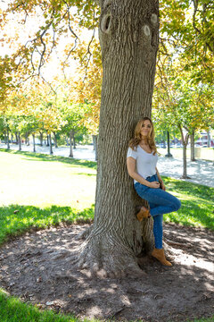 Portrait of a smiling woman in t-shirt and jeans standing in a park leaning against a tree with her knee raised