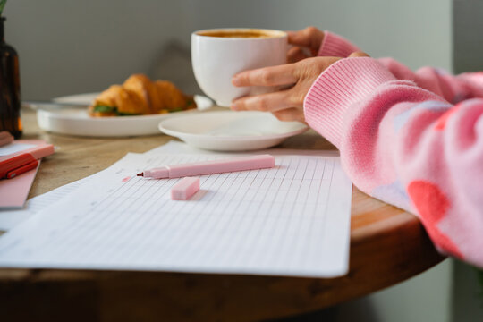 Side view of a woman sitting at a table with a weekly planner drinking a cappuccino and eating a savoury croissant