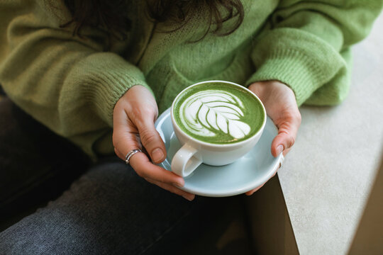 Close-up of a woman in a green cardigan sitting in a cafe holding a cup of hot matcha latte
