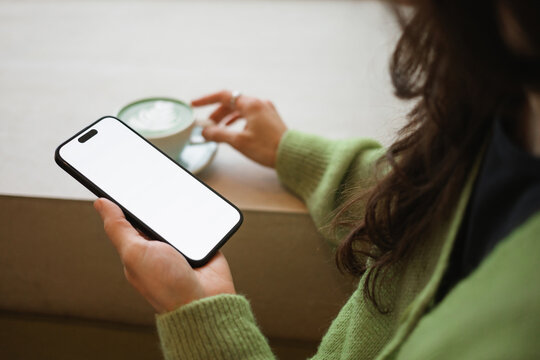 Close-up side view of a young woman drinking a hot matcha latte while holding a mobile phone with a blank screen