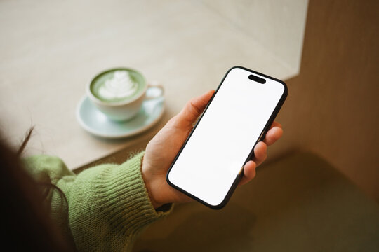 Side view of a young woman drinking a hot matcha latte while using a mobile phone with a blank screen