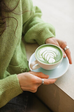 Close-up side view of a woman in a green cardigan sitting at a table in a cafe drinking a hot matcha latte drink