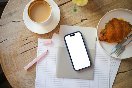 Overhead view of a mobile phone with a blank screen on a weekly planner with a highlighter pen, cup of coffee, savoury cheese and salad croissant and bunch of flowers on a wooden table