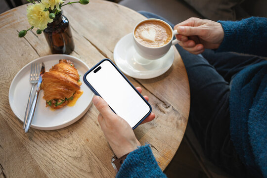Close-up of a man sitting at a table with a cappuccino and savoury croissant holding a mobile phone with a blank screen
