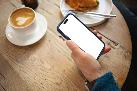 Close-up of a man sitting at a table with a cappuccino and savoury croissant holding a mobile phone with a blank screen
