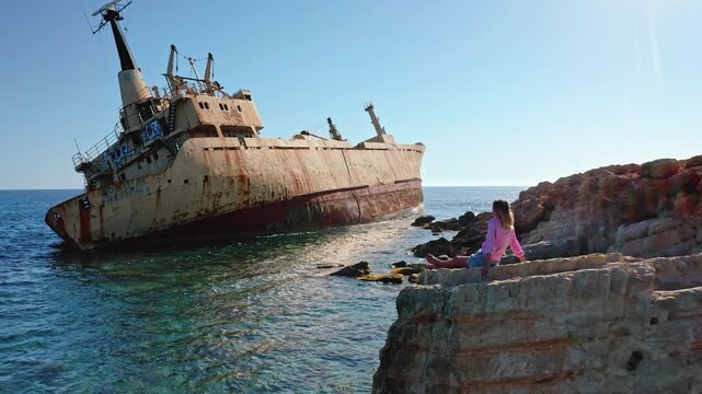 Young woman sitting on a rocky seashore admiring the iconic Edro III shipwreck, a popular tourist landmark near Paphos, under a bright blue sky on a clear day