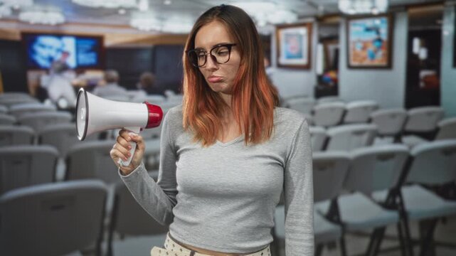 Woman holding megaphone in conference seating building, wearing glasses and sweater, single hand grip, slight slouch and neutral expression; boredom.