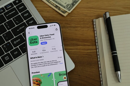 Kiel, Germany - 02-25-2026: A smartphone displaying the Uber Eats food delivery and restaurant ordering app in App Store displayed on smartphone placed on top of laptop on wooden desk