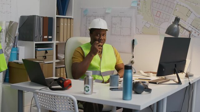 Medium long shot of African American male construction engineer in safety vest and hardhat using walkie-talkie while working at office desk with computer and building plans