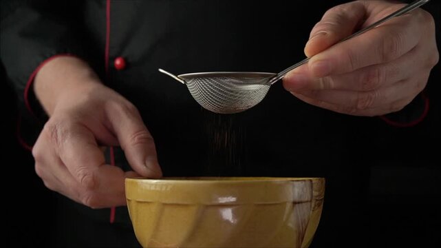 Chef's hands using a fine mesh strainer to sift cocoa powder into a yellow bowl, showcasing a precise cooking technique in a dark kitchen setting
