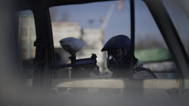 A paintball player in full gear holds a paintball gun, looking out from a car window. The participant surveys the field, ready for action during the game.