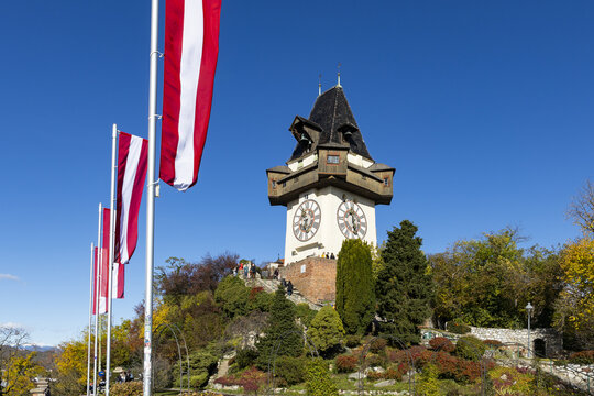 View of the clock tower standing tall on a hill with flags waving in the wind against a blue sky, Graz, Styria, Austria.