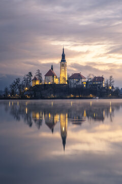 View of a serene island church illuminated against a twilight sky, mirrored perfectly on the tranquil lake's surface, creating a magical scene, Bled, Radovljica, Slovenia.