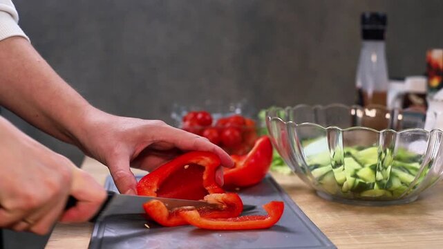 Caucasian person slices red bell pepper on a cutting board with fresh vegetables and condiments visible in the kitchen, showcasing the preparation of a healthy meal in a home setting