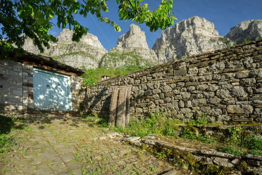 View of a stone wall bordering a weathered building under the towering presence of the mountains, contrasting the old with the sublime, Mikro Papigko, Ioannina, Greece.