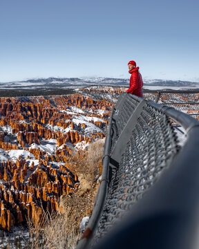 View of a person in bright red jacket stands on a metal overlook, gazing at the snow-dusted orange hoodoos stretching across the frosted landscape, Bryce Canyon City, Utah, United States.