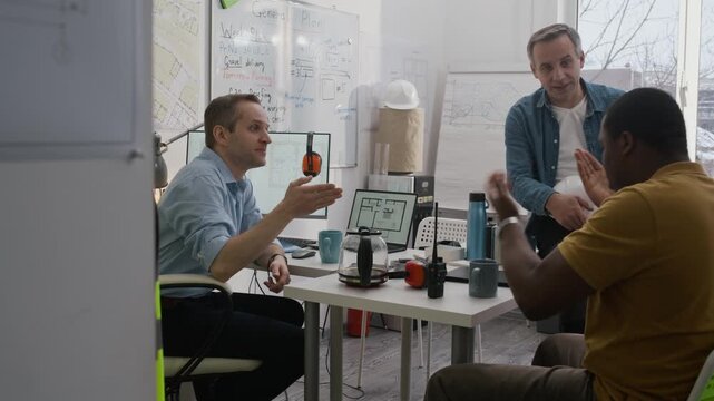 Medium long shot of three diverse male civil engineers having tense work conversation while meeting in construction office