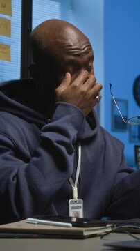 Vertical shot of exhausted Black millennial programmer dressed in hoodie with badge rubbing tired eyes during workday at office desk
