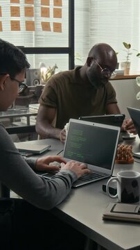 Vertical shot of two diverse web engineers programming at wooden desks during workday in modern office of tech company