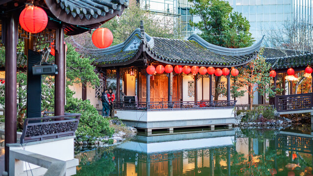 View of a serene Chinese garden pavilion reflected in still water, adorned with vibrant red lanterns against a backdrop of lush greenery, Portland, Oregon, United States.