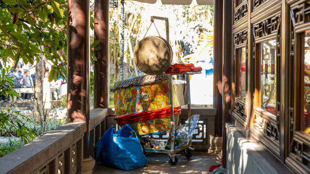 View of a gong hanging above a cart with vibrant fabric and a blue bag, framed by ornate wooden architecture and lush greenery, Portland, Oregon, United States.