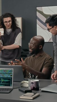Vertical shot of positive Black young IT specialist sharing idea with two Caucasian gen Z programmers while sitting at his desk in front of laptop screen