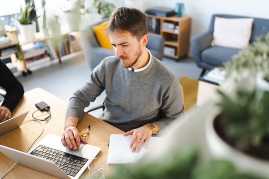 A man works on his laptop and takes notes in a notebook at a wooden desk in a bright, modern office space.