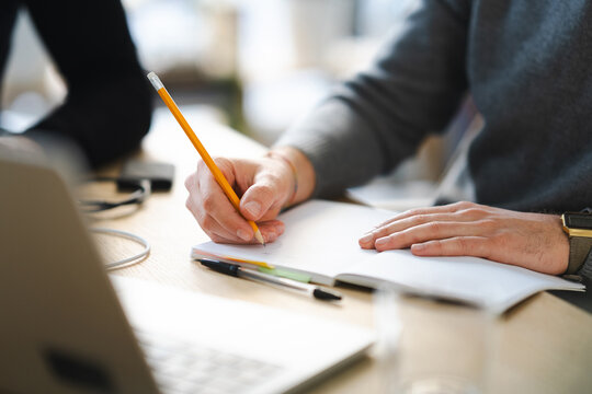 A person's hands are shown writing in a notebook with a pencil, with a laptop and pens on the desk.