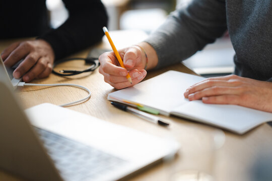 Two people collaborate at a desk, one typing on a laptop while the other writes in a notebook with a pencil.