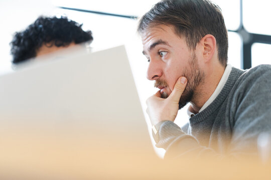 A man with a beard looks intently at a computer screen, his hand resting on his chin in thought. Another person is partially visible in the background.