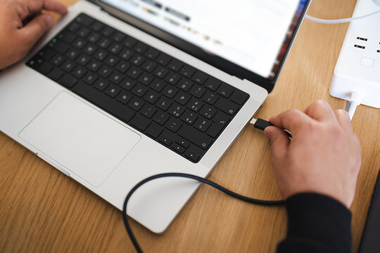 A person connects a charging cable to a laptop, with a power strip visible in the background, on a wooden desk.