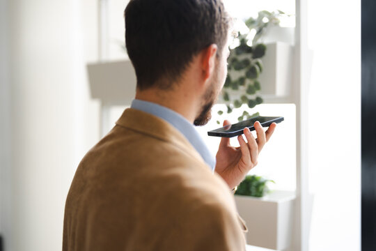 A man in a tan jacket speaks into his smartphone, using its voice assistant feature.