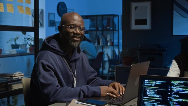 Portrait of Black millennial QA engineer wearing hoodie smiling at camera while testing software product in dimly lit office