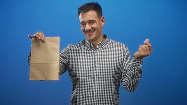 Young hispanic man holds a brown paper bag with right hand and beckons with left hand in studio; playful offer.