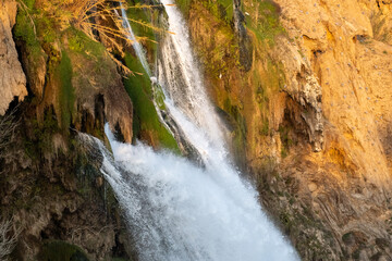 A detailed close-up photograph capturing the powerful flow of the Lower Duden Waterfall as it cascades over rugged, brown rocky cliffs. The image highlights the vibrant green moss covering the wet roc © osmanmaasoglu