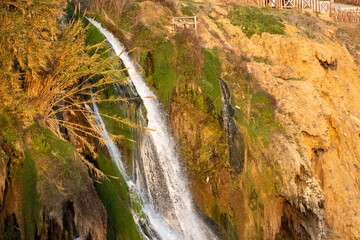 A detailed close-up photograph capturing the powerful flow of the Lower Duden Waterfall as it cascades over rugged, brown rocky cliffs. The image highlights the vibrant green moss covering the wet roc © osmanmaasoglu