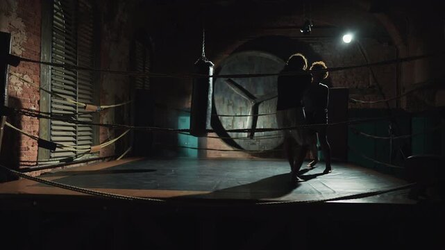 African man and woman training on boxing ring, full-length shot in dark gym. Sport workout for developing endurance and force, self-defense class for women, training punches by arms, box workout