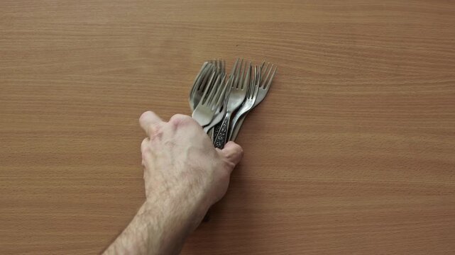 A person hands choosing several shiny metallic forks featuring different handle patterns from a jumbled pile of cutlery, resting on natural wood grain desktop, illustrating kitchen chore