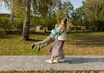 Fototapeta premium An older sister spins her younger brother around, holding his armpits and spinning around in the park. Children are having fun outdoors, enjoying the moment of joy. Life energy. Dynamic photo