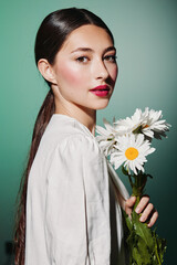 Young woman with glossy dark hair and bright makeup holds white daisies in hand against blue...