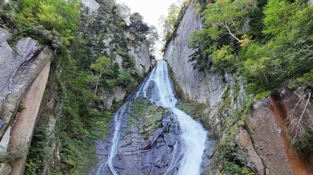 Dynamic low-angle aerial footage looking up at Ginga Waterfall in Sounkyo Gorge, Hokkaido, Japan. The video captures the majestic scale of the waterfall cascading down the steep columnar jointing clif