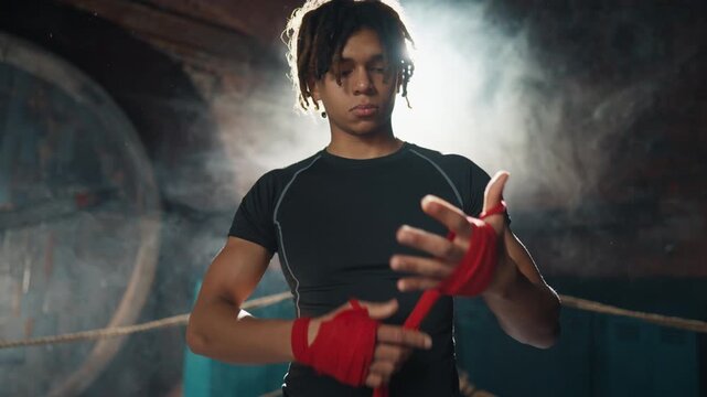 Handsome young african man wrapping protective tape on hand before training. Professional boxer fighter preparing to fight or workout, cinematic portrait in dark gym, do sport for keep fit, be strong