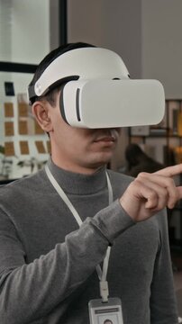 Vertical shot of gen Z male web designer with badge developing game using VR headset in modern office