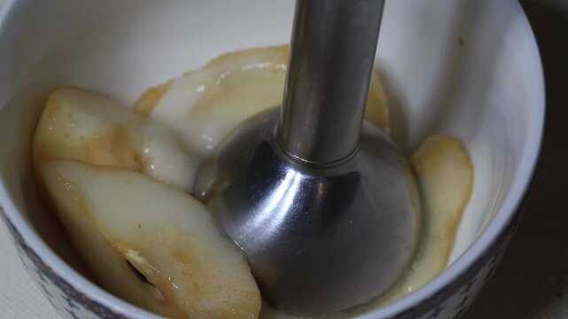 Close-up view of immersion blender operating within ceramic bowl, transforming soft cooked fruit into a smooth, creamy puree. Culinary process involves blending warm, tender fruit slices to create a w