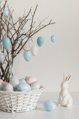 Obraz premium Easter tree with pastel speckled eggs hanging on spring branches in glass vase. White ceramic bunny and basket with painted eggs on light background. Soft natural light, minimal home decor