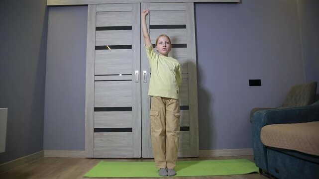 Full body shot of young boy performing arm exercises indoors. Child stands on yoga mat, raising and lowering arms in rhythmic motion, demonstrating physical activity and home fitness routine.