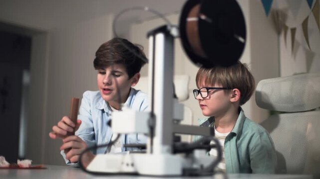 Two children at a table with a working 3D printer, discussing a printed part