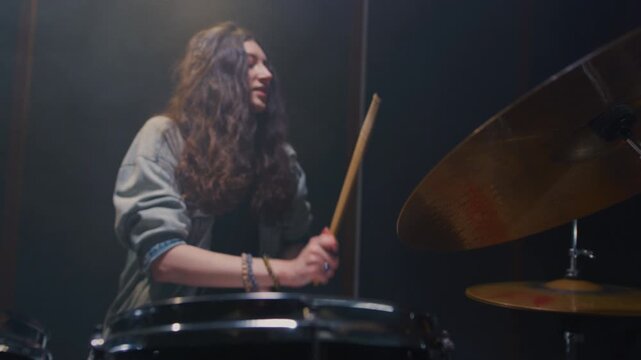 Portrait: female with long curly hair plays the drums. Girl is rehearsing in a studio with drumsticks. Face of a beautiful musician girl, showcasing skill and energy. Underground indie slow motion