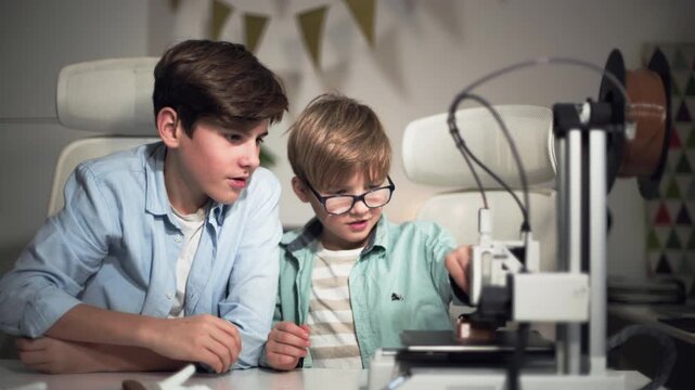 At a bright table, two schoolchildren watch a 3D printer work and play with it